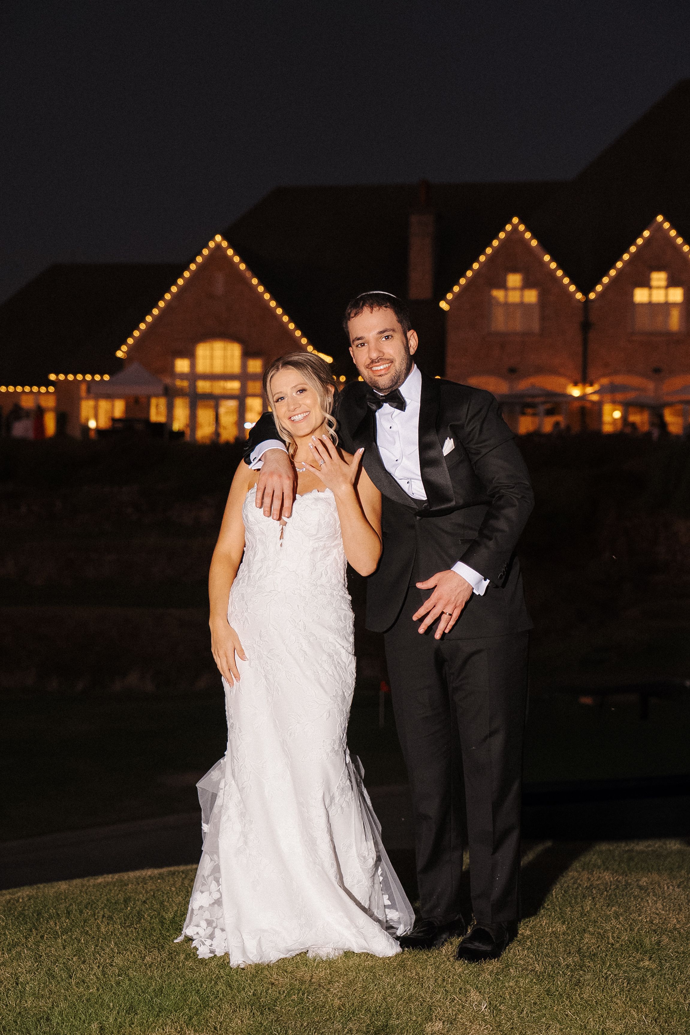 A couple poses playfully on a lawn at night with illuminated houses glowing warmly in the background.