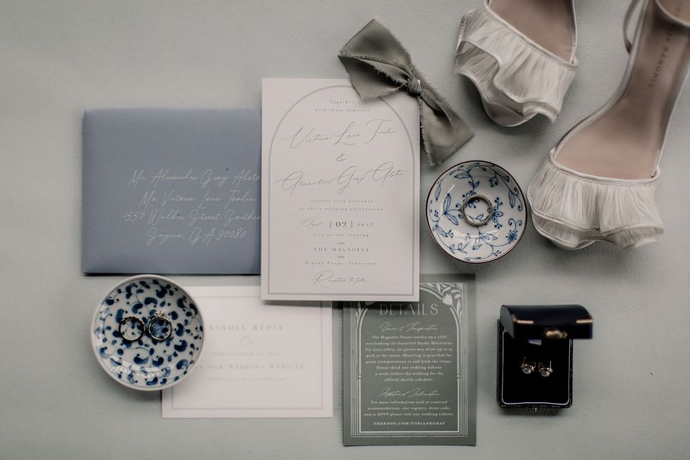 Elegant wedding invitation suite with soft pink and gray tones featuring rings and floral details laid out on white surface.