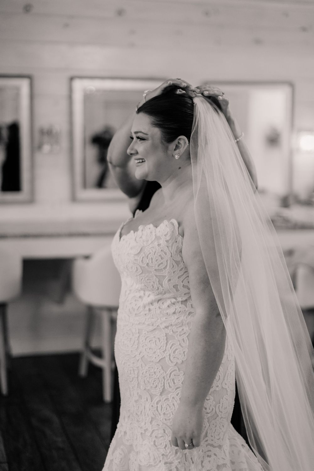 A bride in a detailed lace wedding dress smiles joyfully while wearing a long flowing veil in a bridal suite.
