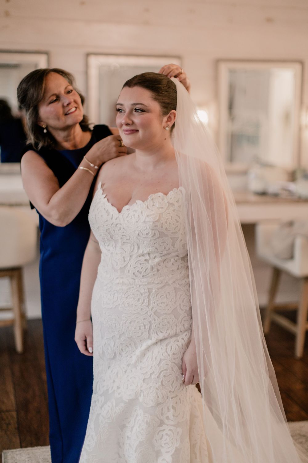A bride in a lace wedding dress gets help with final preparations in a bright indoor setting.