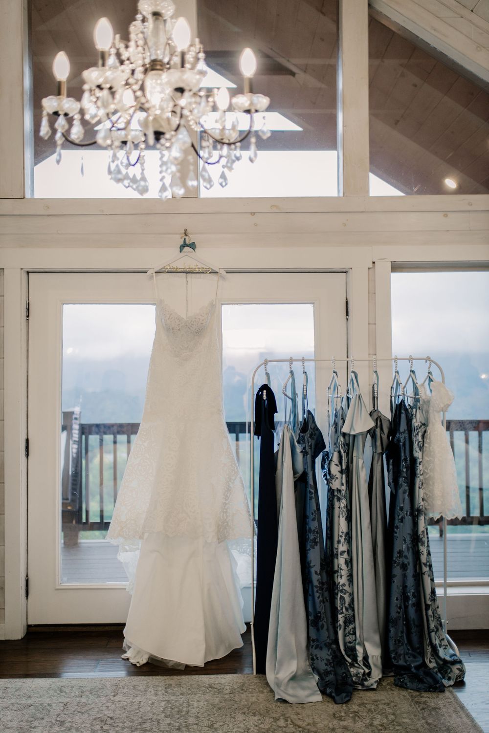 Wedding dresses and bridesmaid gowns hanging from chandeliers in an elegant venue with large windows.