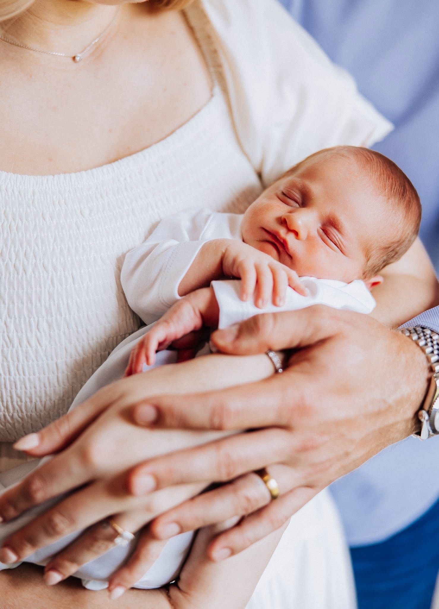 Sleeping newborn baby cradled in parents hands, mother wearing a white dress and gold ring.
