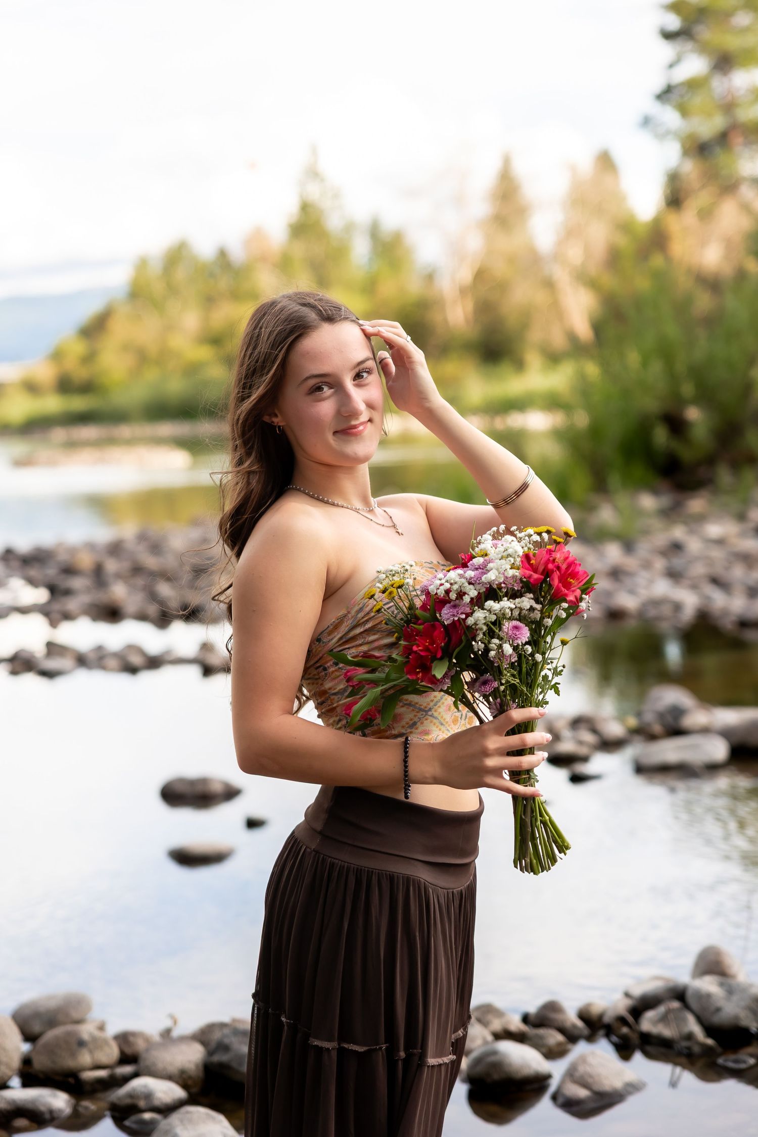 Person in brown dress poses with flowers by rocky lakeshore at sunset in outdoor portrait series.