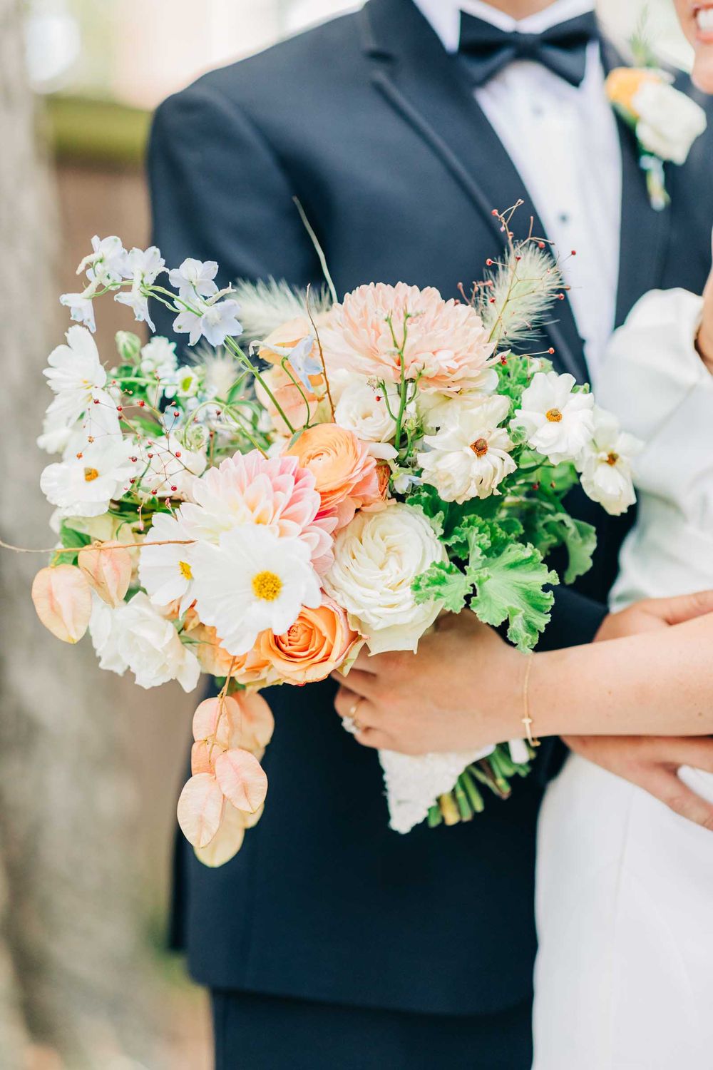 A beautiful wedding bouquet with peach, white and blush flowers held against a navy blue suit at 255 Milledge, Hardeman - Sams Estate