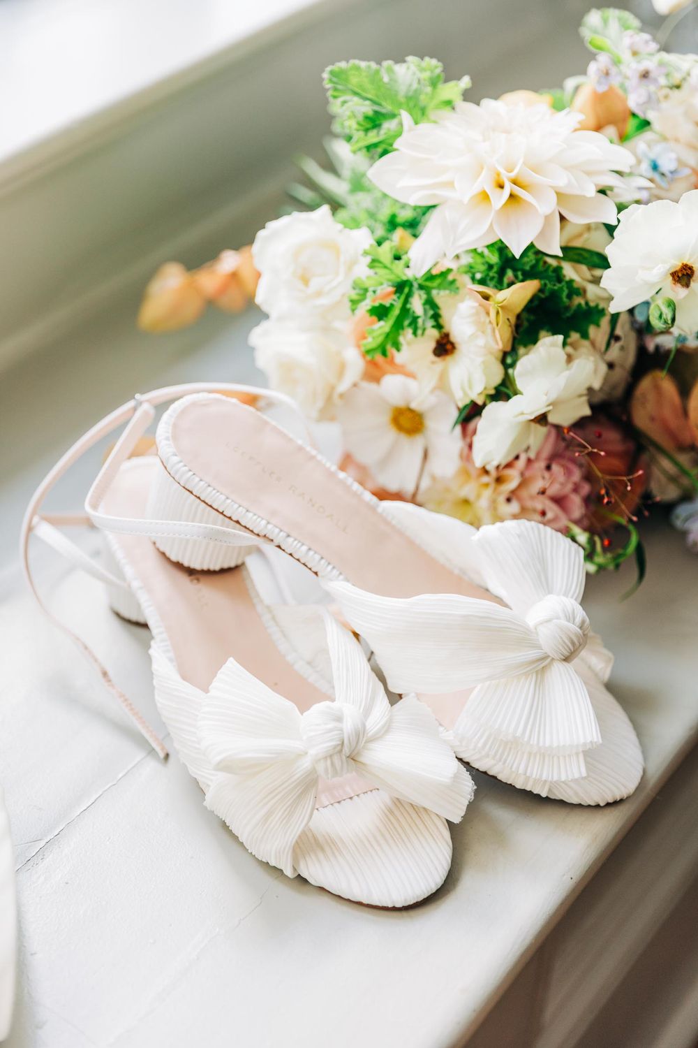 Elegant bridal heels with bow next to a soft pastel floral bouquet on a light surface at 255 Milledge, Hardeman - Sams Estate
