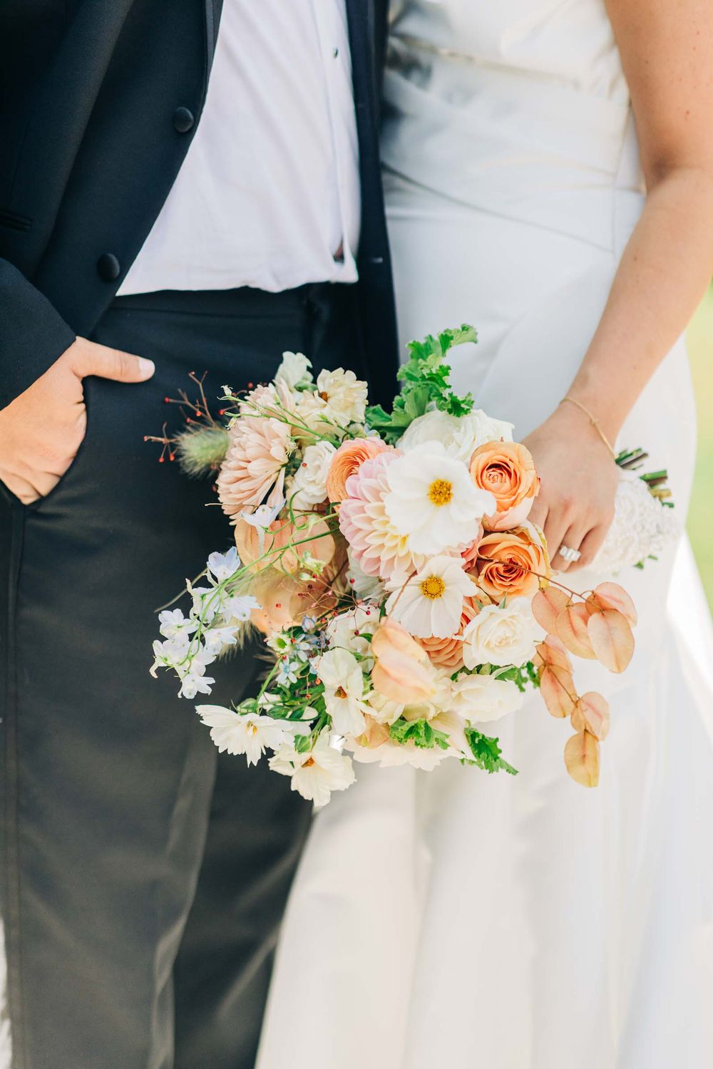Delicate peach and white wedding bouquet with roses and greenery held against a white dress at 255 Milledge, Hardeman - Sams Estate
