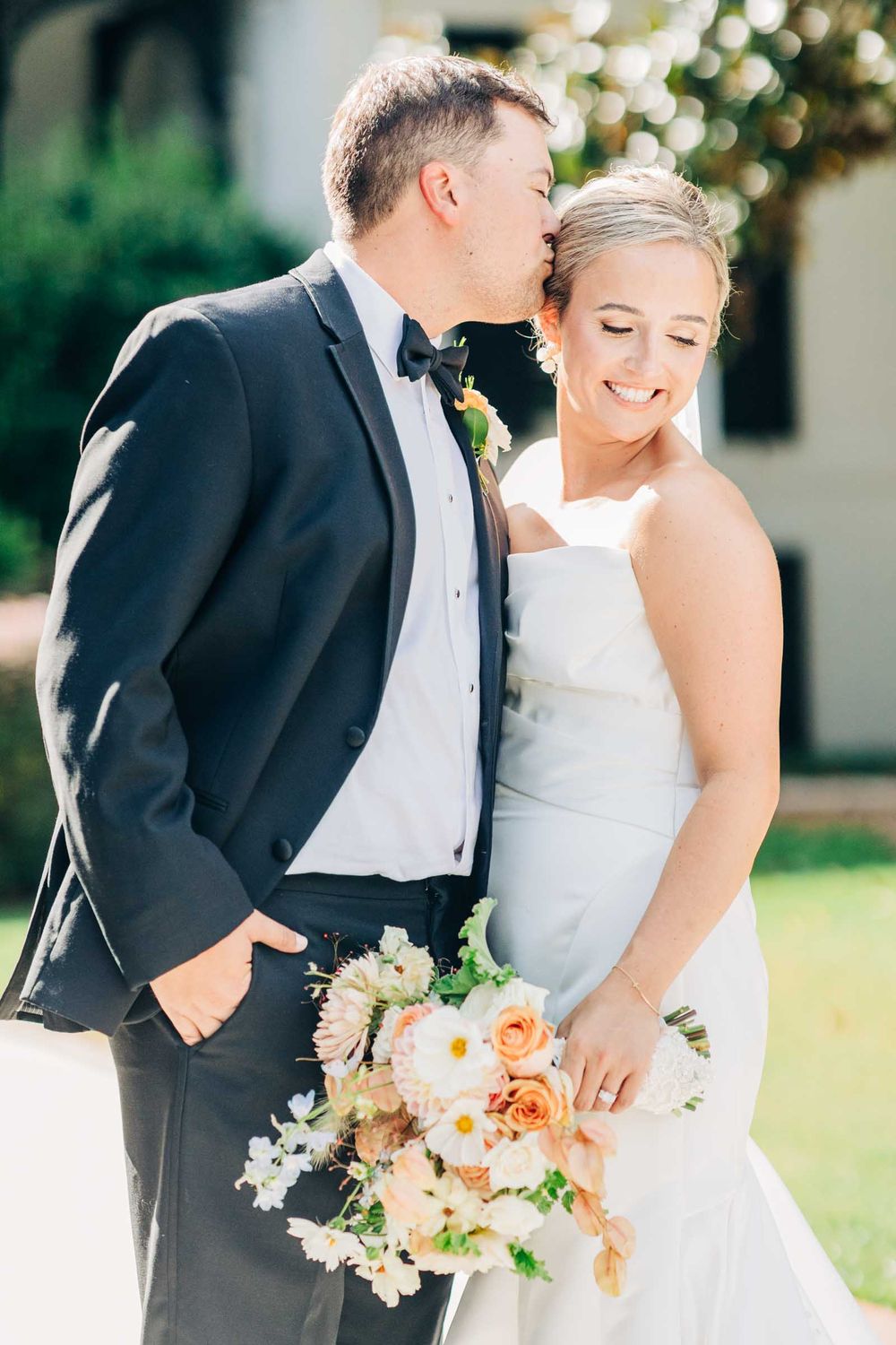 A romantic wedding couple embraces outdoors while holding a peach and cream floral bouquet at 255 Milledge, Hardeman - Sams Estate