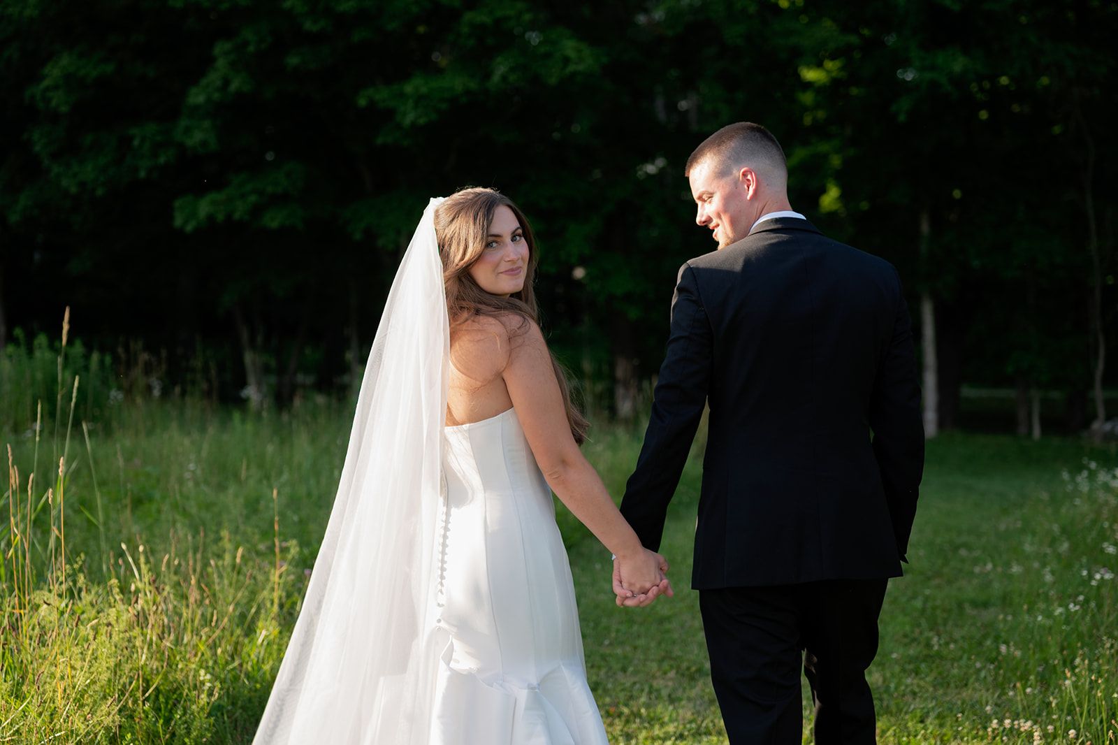 A couple in wedding attire holds hands while walking through a lush green meadow on their special day.