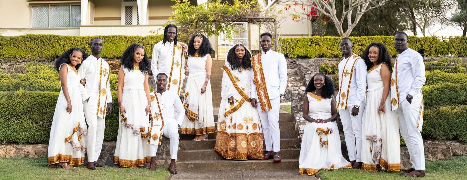 Group of people in matching white and gold traditional Ethiopian wedding attire posing on outdoor steps.