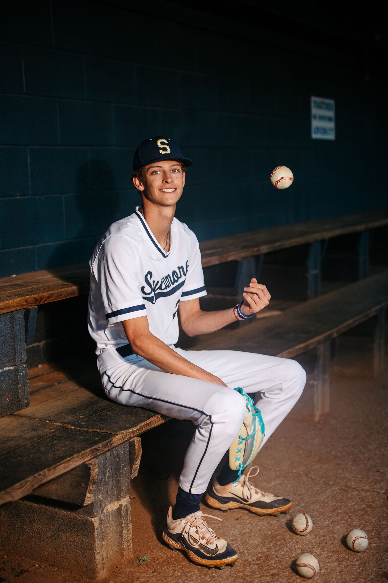 Baseball player in white uniform sits in dugout tossing a ball while surrounded by baseballs on wooden bench.