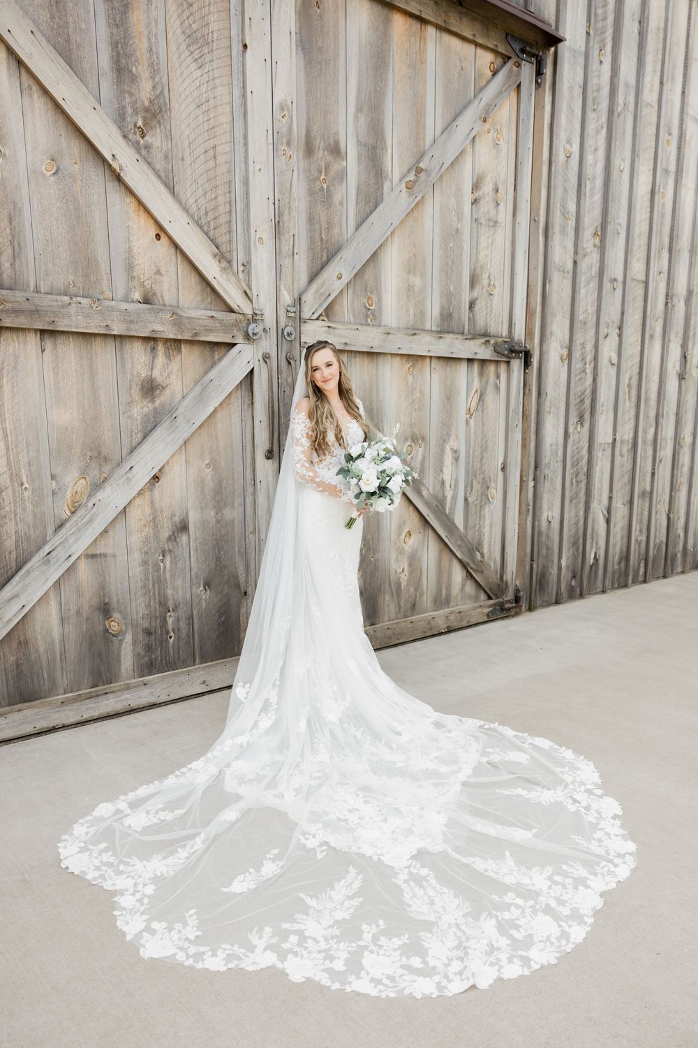 Stunning cathedral length lace train of white wedding gown against wooden barn wall.