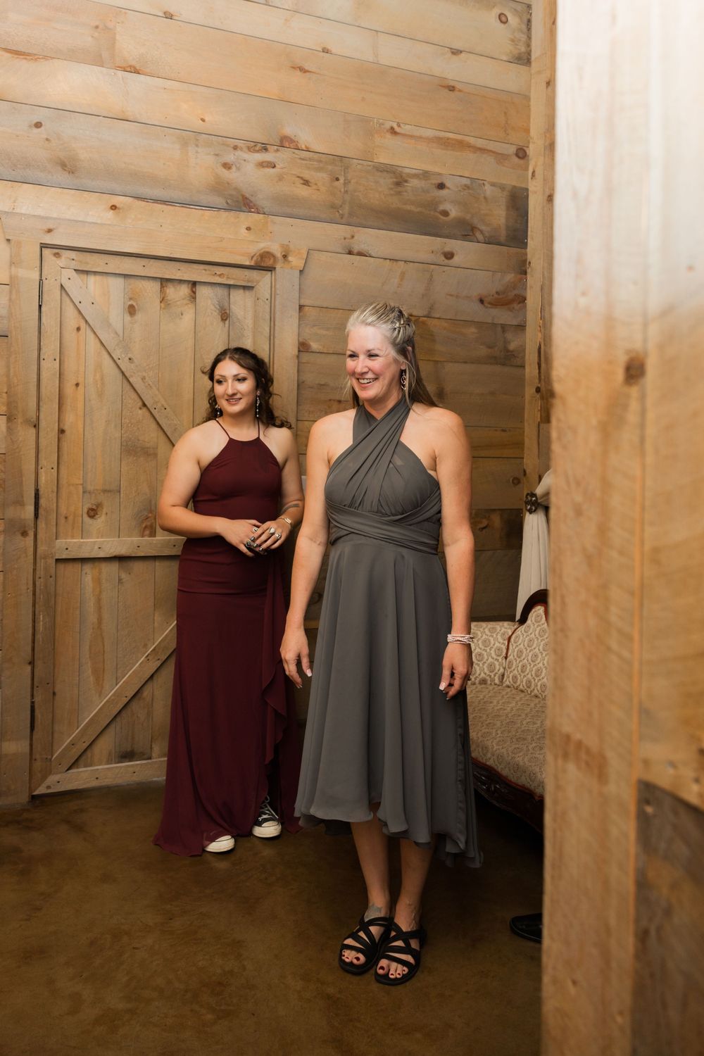 Wedding party members in burgundy and gray dresses pose inside a rustic barn venue.