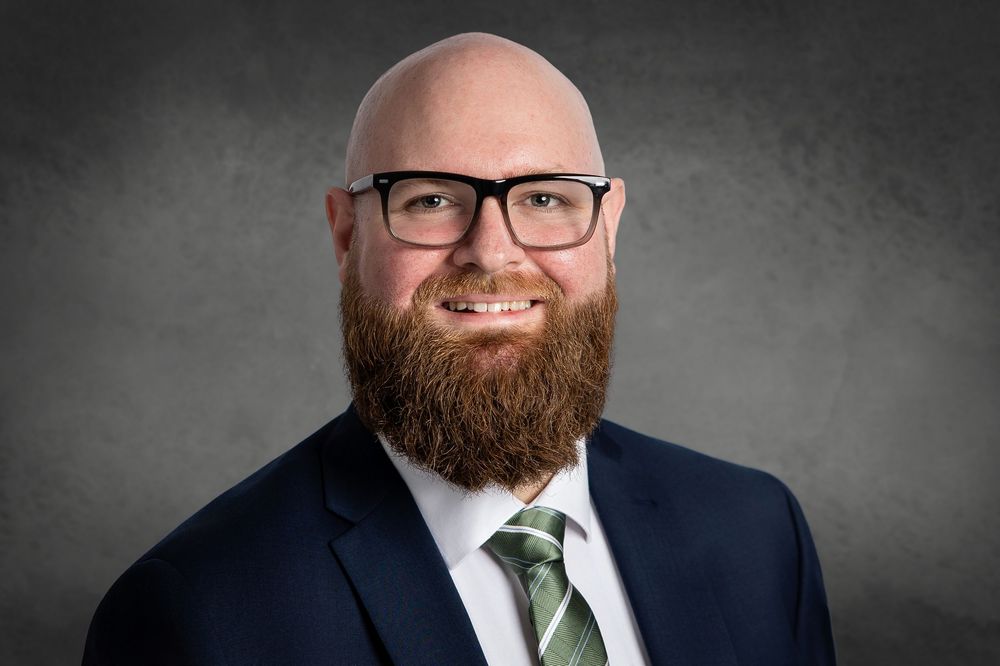 Professional headshot of person in navy suit and green tie wearing glasses with full beard against gray background.
