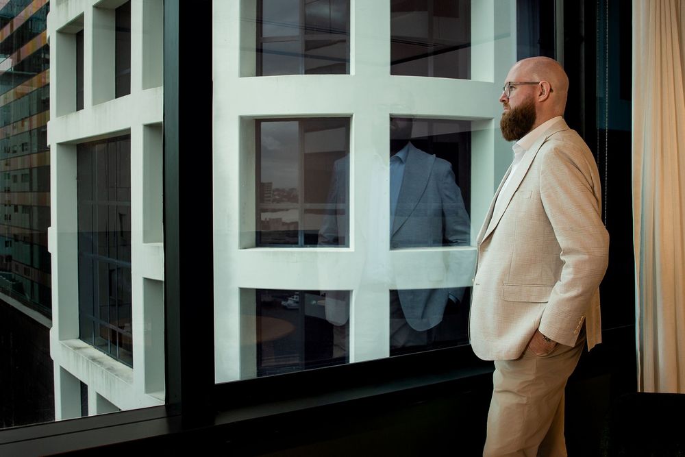 A person in a cream-colored suit stands contemplatively by large geometric windows of a modern building.