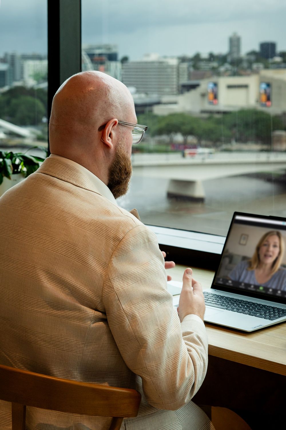 A person in a beige jacket participates in a video conference call at a desk with a laptop and city view.