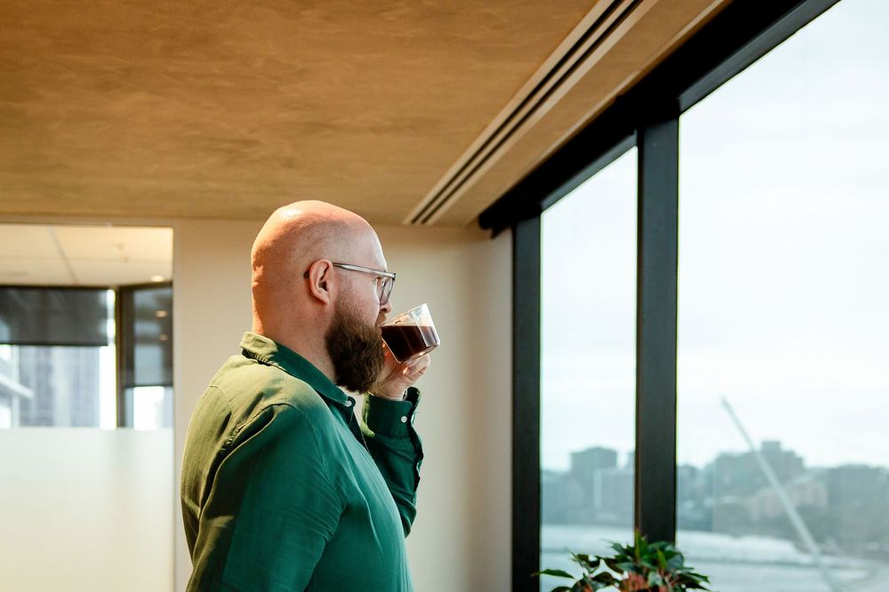 Person in bright green sweater drinks from a mug while looking out large office window.