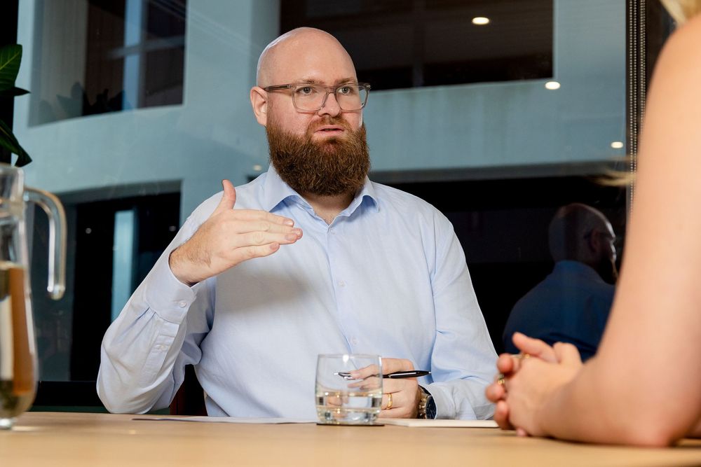 A person with a beard and glasses sits at a restaurant table holding a glass of whiskey.