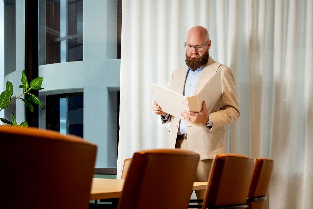 Business professional in a tan suit checks his watch while standing near large windows in a modern office setting.