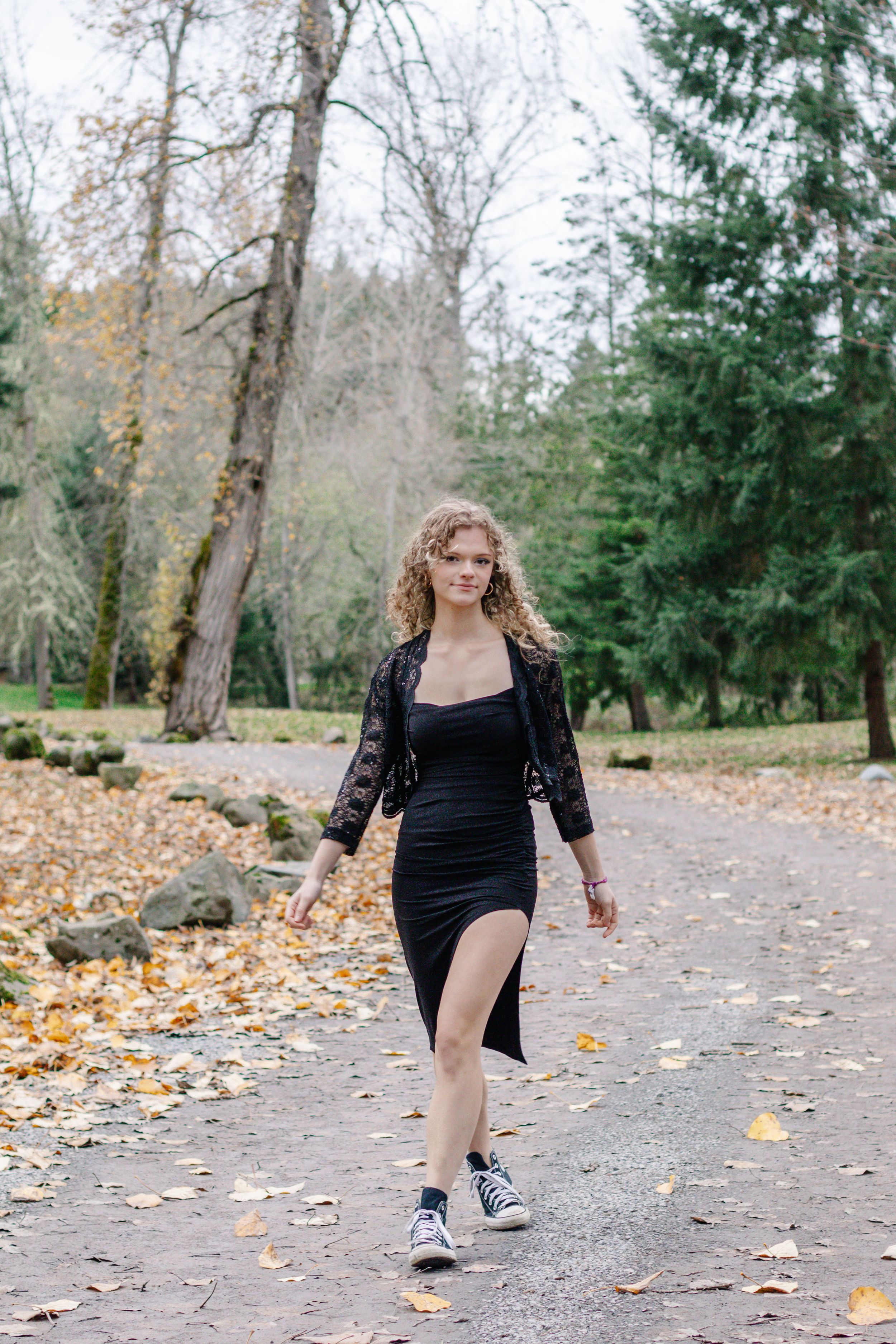 Young woman in black dress and sneakers walking on leaf-covered park path.