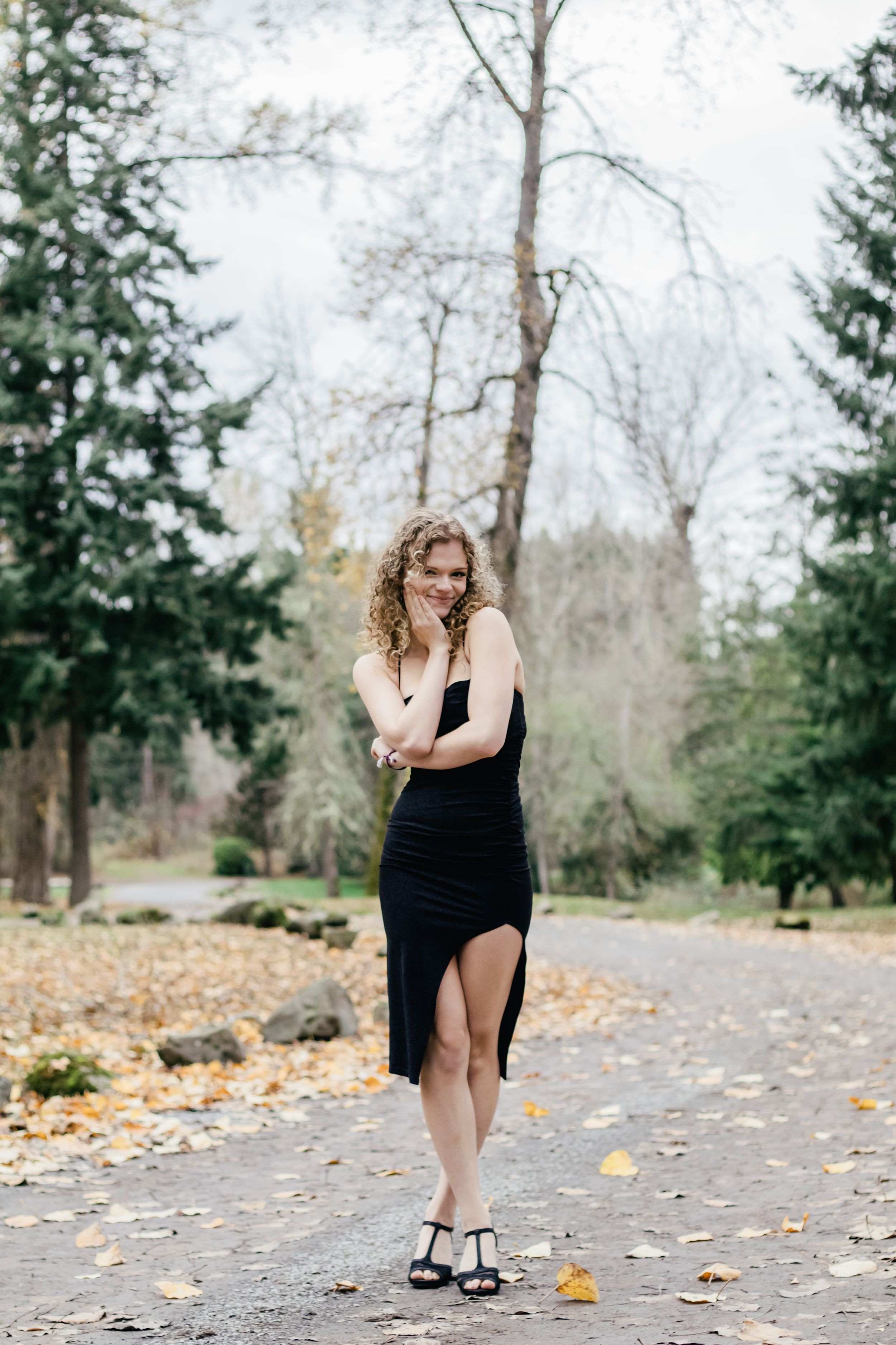 Curly-haired young woman in black dress smiling on a leaf-covered park path.