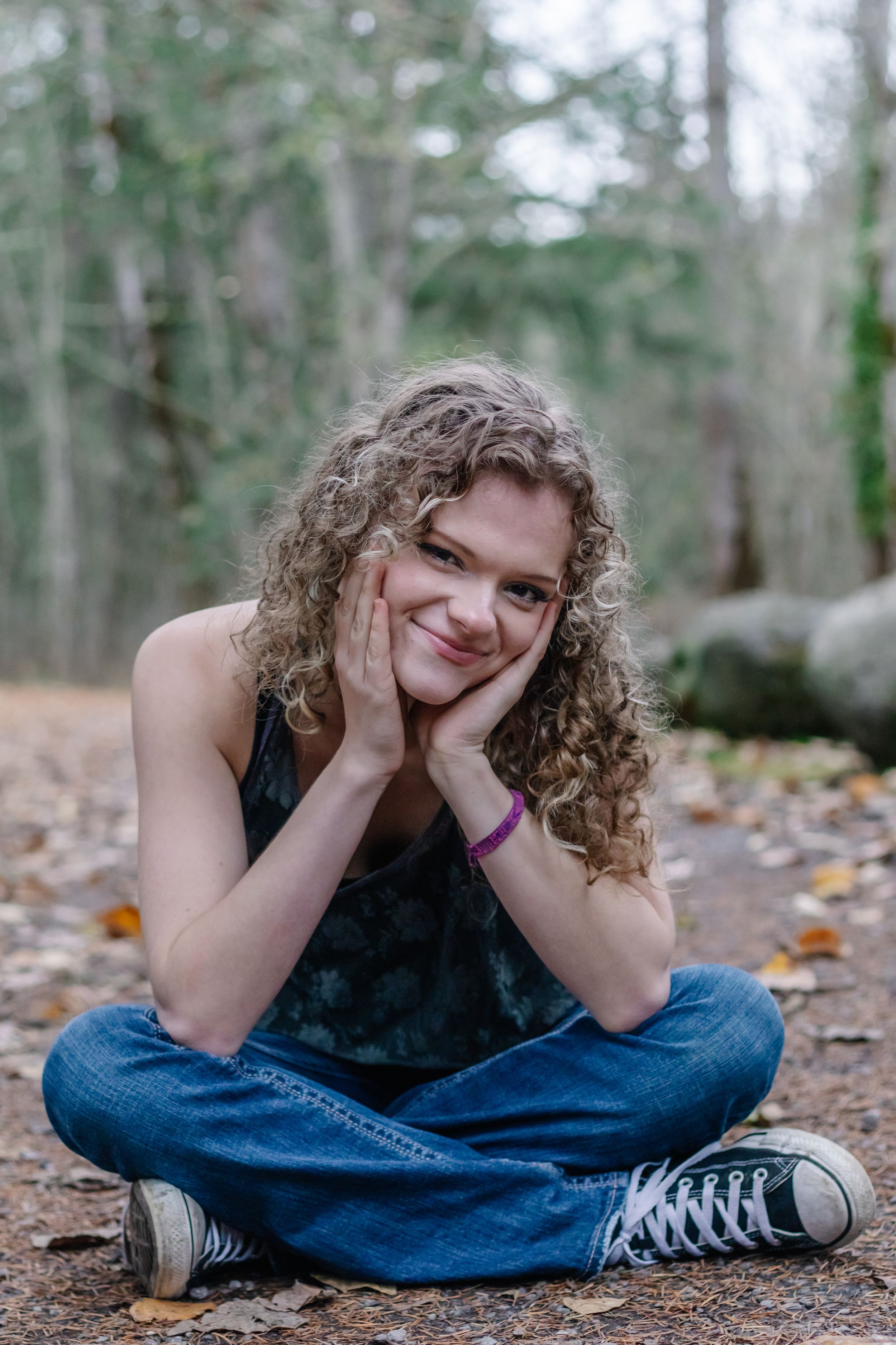 Curly-haired girl sitting cross-legged on forest floor smiling at camera.