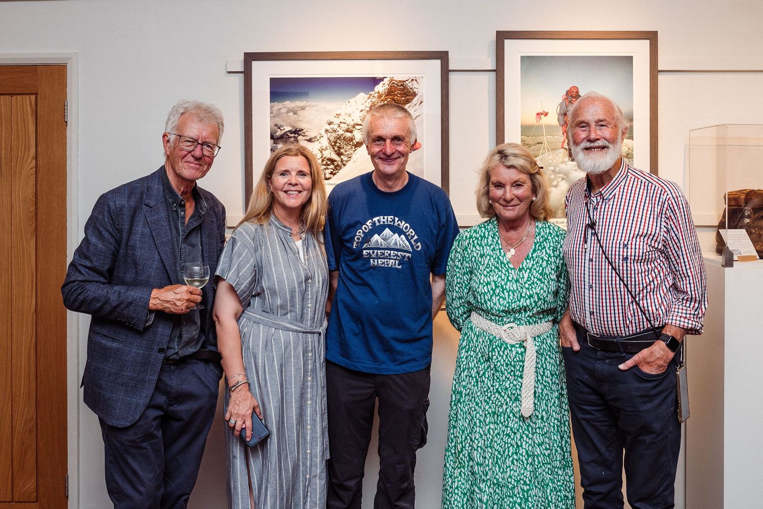 A group of five people standing together indoors in casual attire against a white wall with framed artwork.