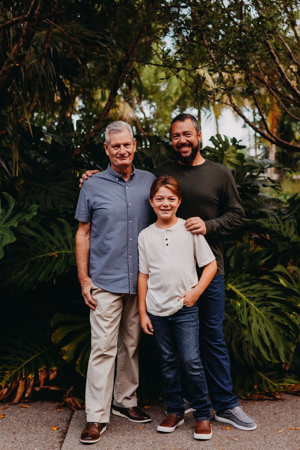 Three people stand together in a casual outdoor setting with lush green plants in the background.