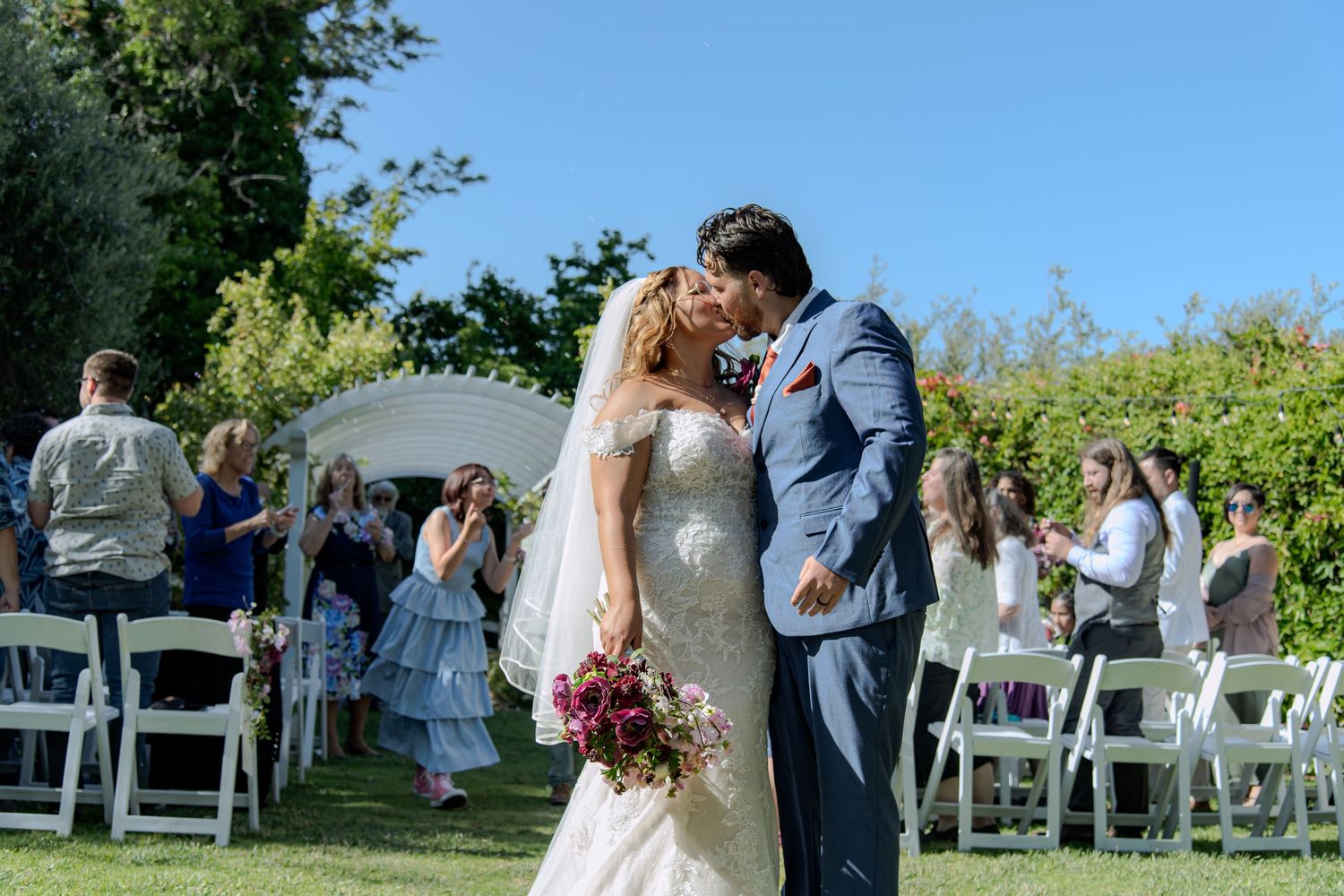 A couple shares a romantic moment during an outdoor wedding ceremony with white chairs and greenery in the background.