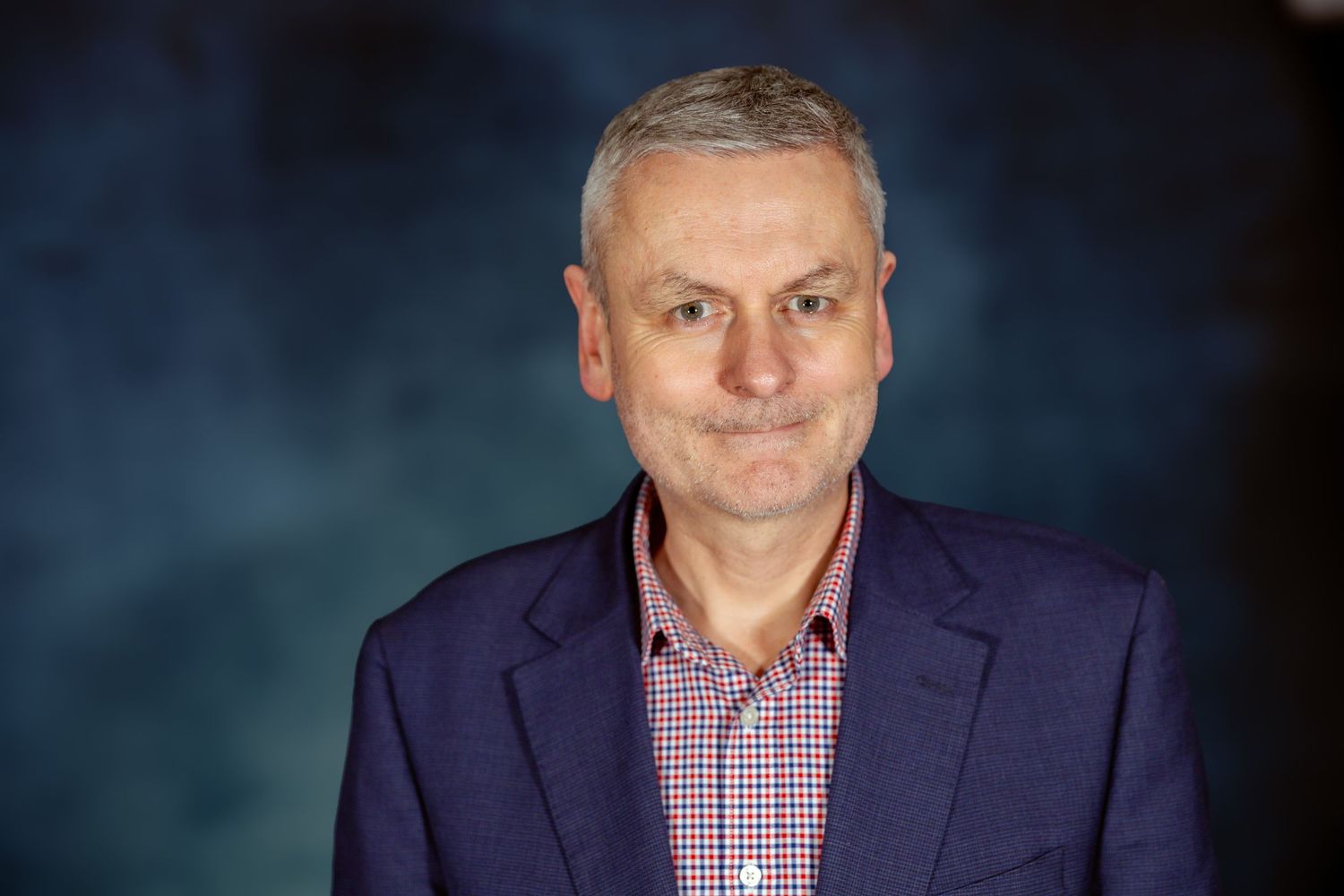 Professional headshot portrait of a person wearing a navy suit jacket and checkered dress shirt against a dark background.