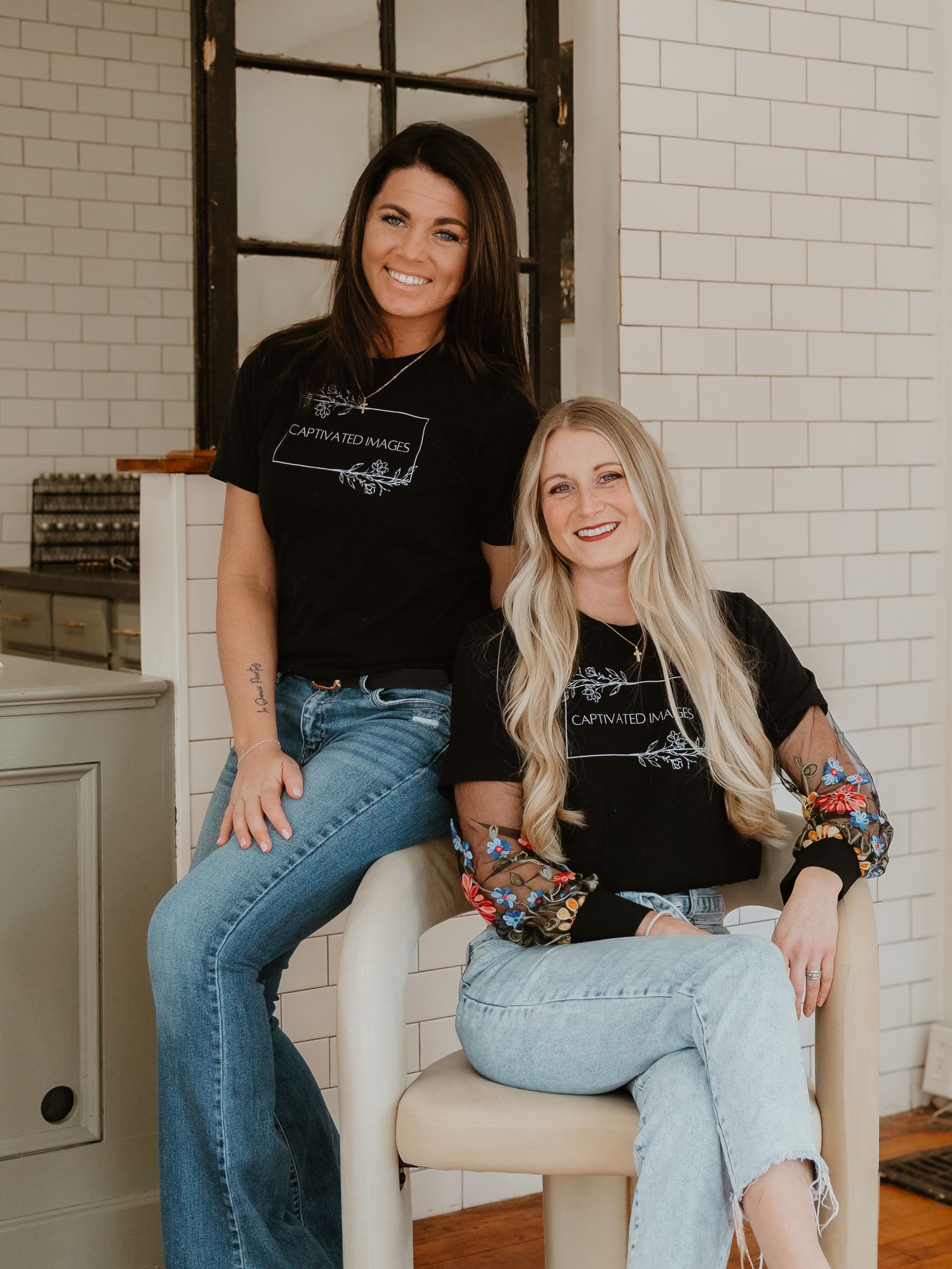 Two friends in matching black t-shirts and blue jeans sit casually on white chairs while sharing a laugh together.