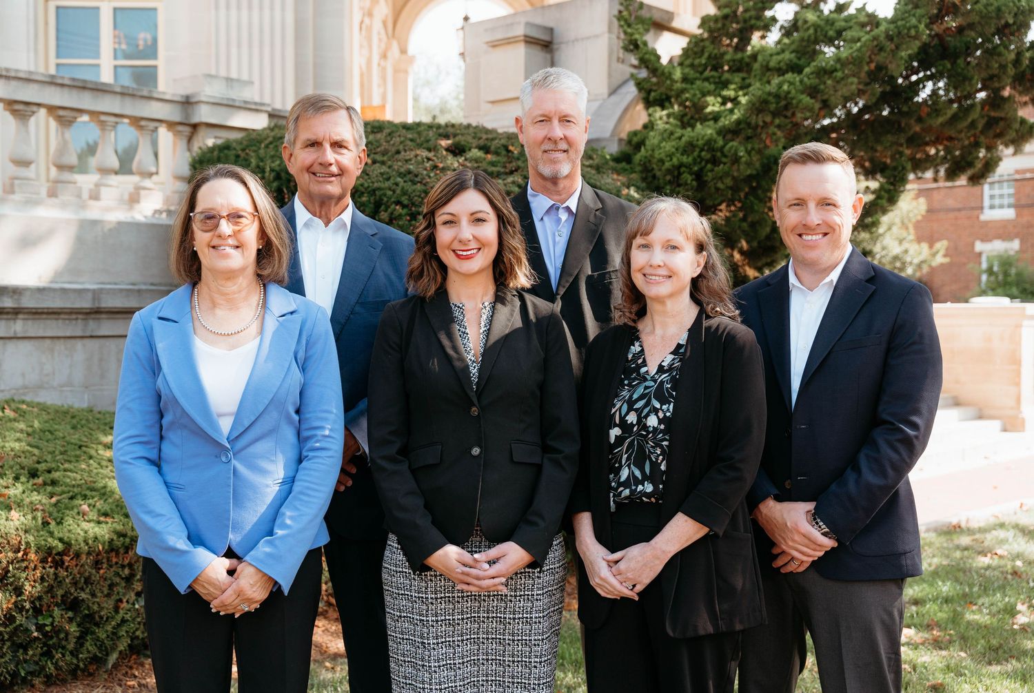 A group of business professionals in formal attire stand together for a corporate team photo outdoors.