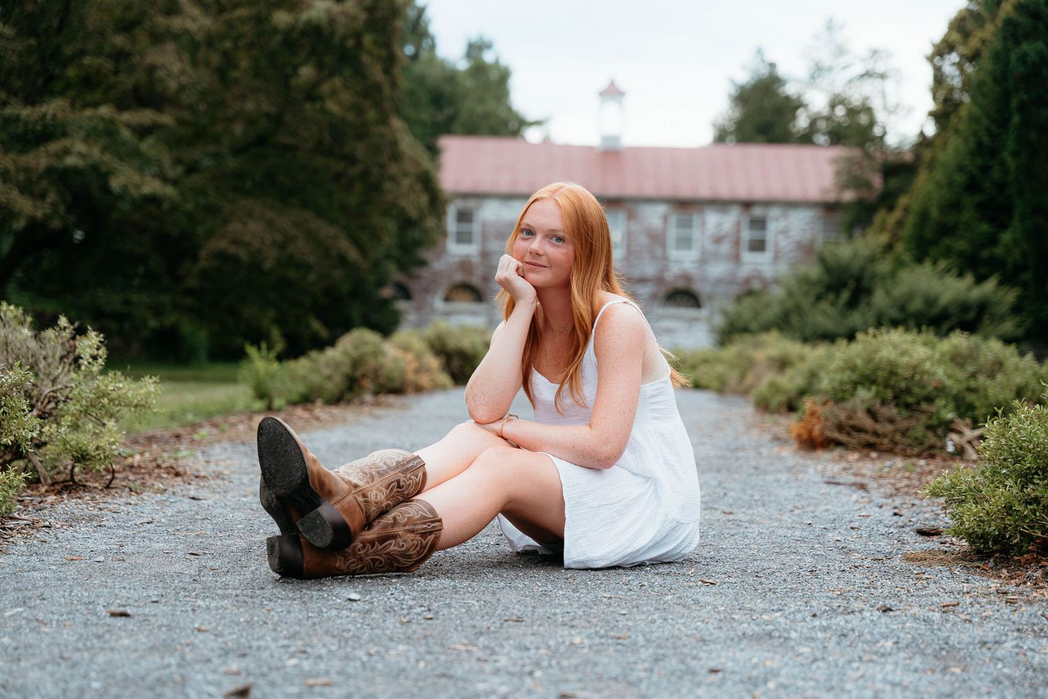 Woman in white dress sitting on gravel path with dog during outdoor photo session.