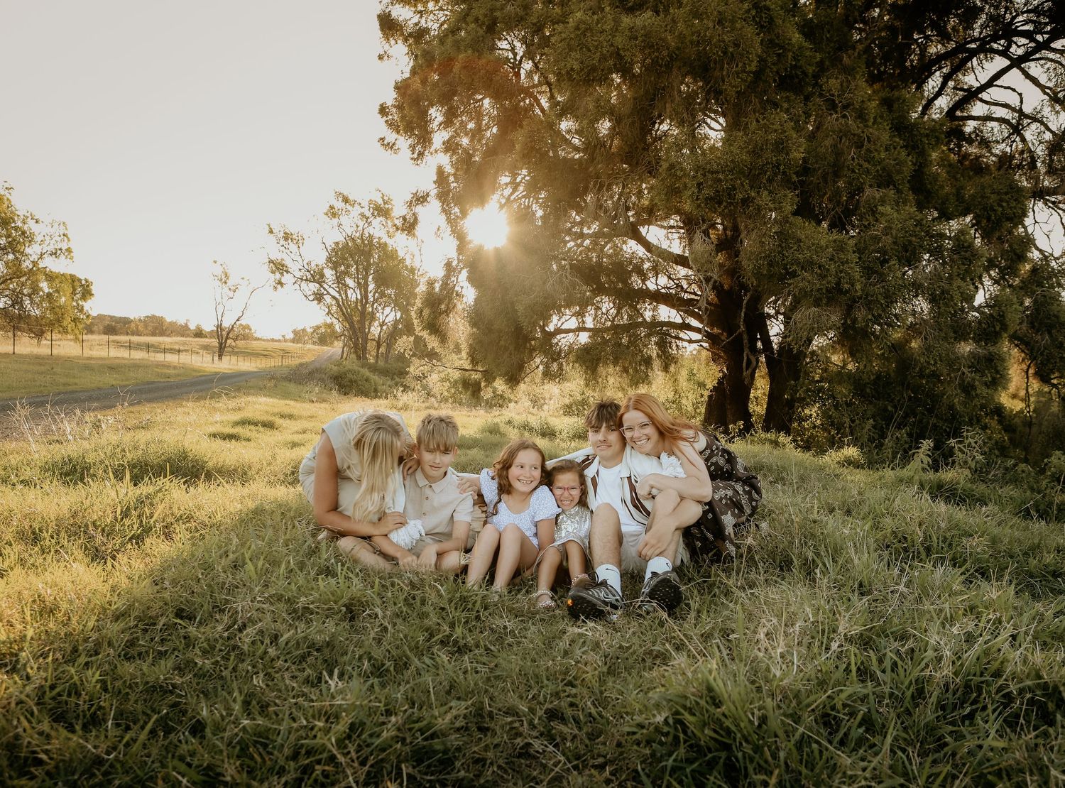 A group sits together on a grassy hillside during golden hour as sunlight streams through a large tree behind them.