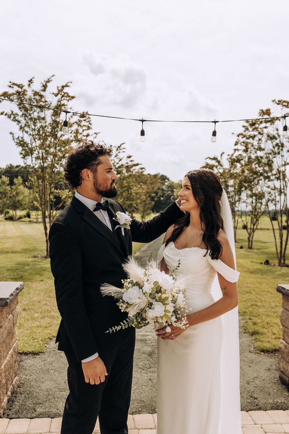 Groom tenderly touching brides veil as she smiles holding a white bouquet at outdoor wedding.