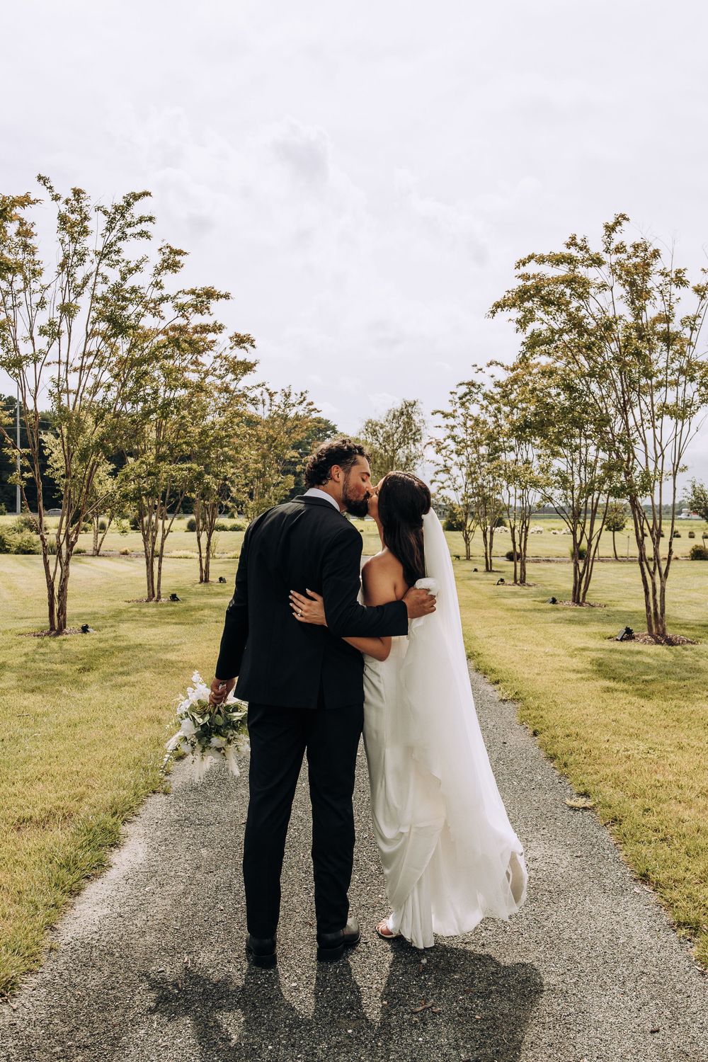 Bride and groom sharing a kiss on a scenic tree-lined pathway at their outdoor wedding.