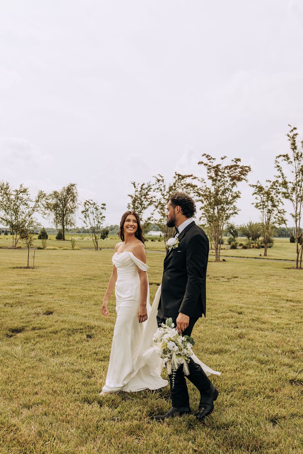 Smiling bride in off-shoulder gown walks with groom carrying bouquet across green lawn.