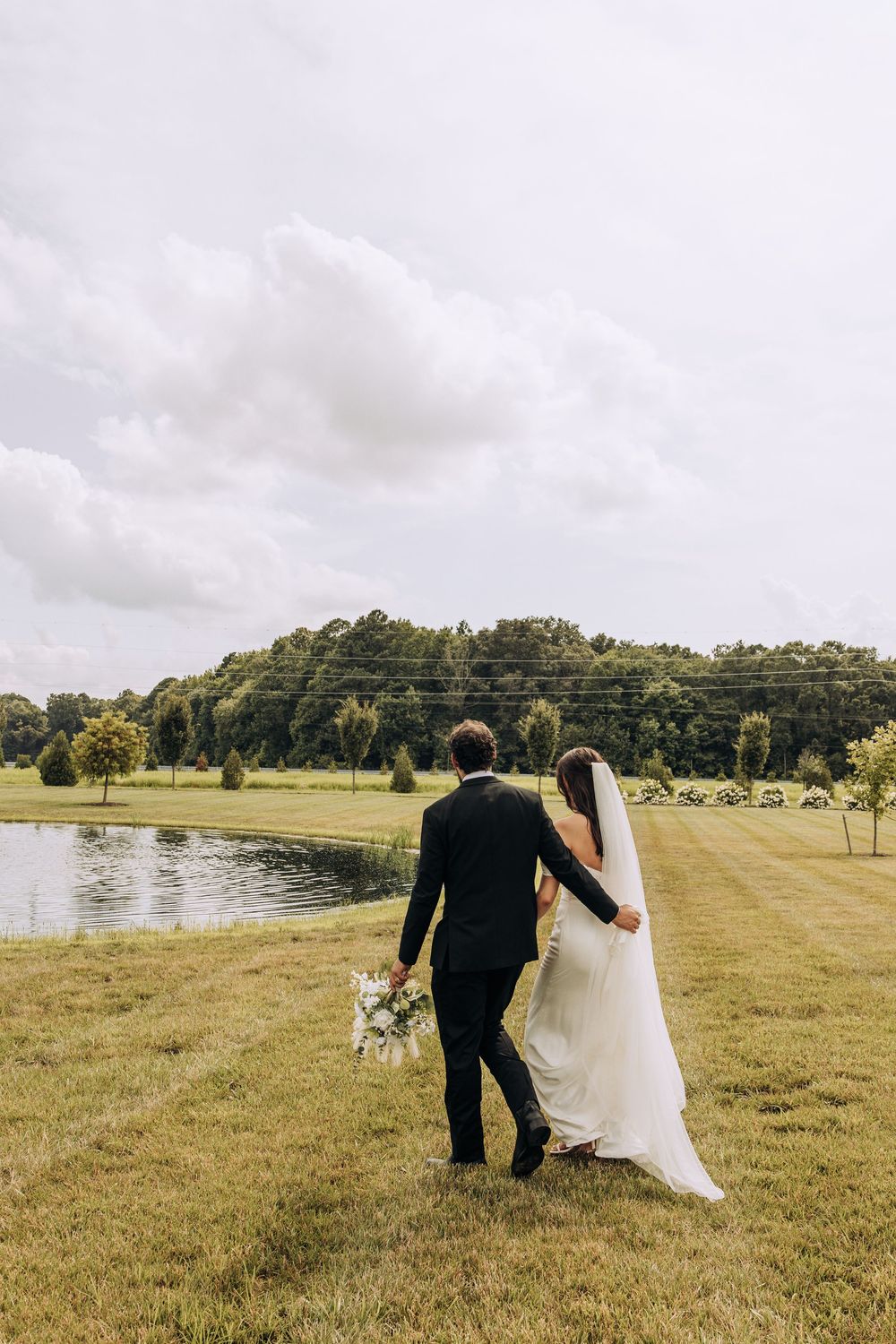 Bride and groom walking arm in arm toward a serene pond at their outdoor wedding venue.