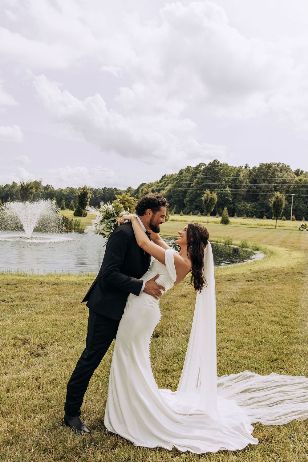 Bride and groom embrace near a pond with fountain on lush green grounds on their wedding day.