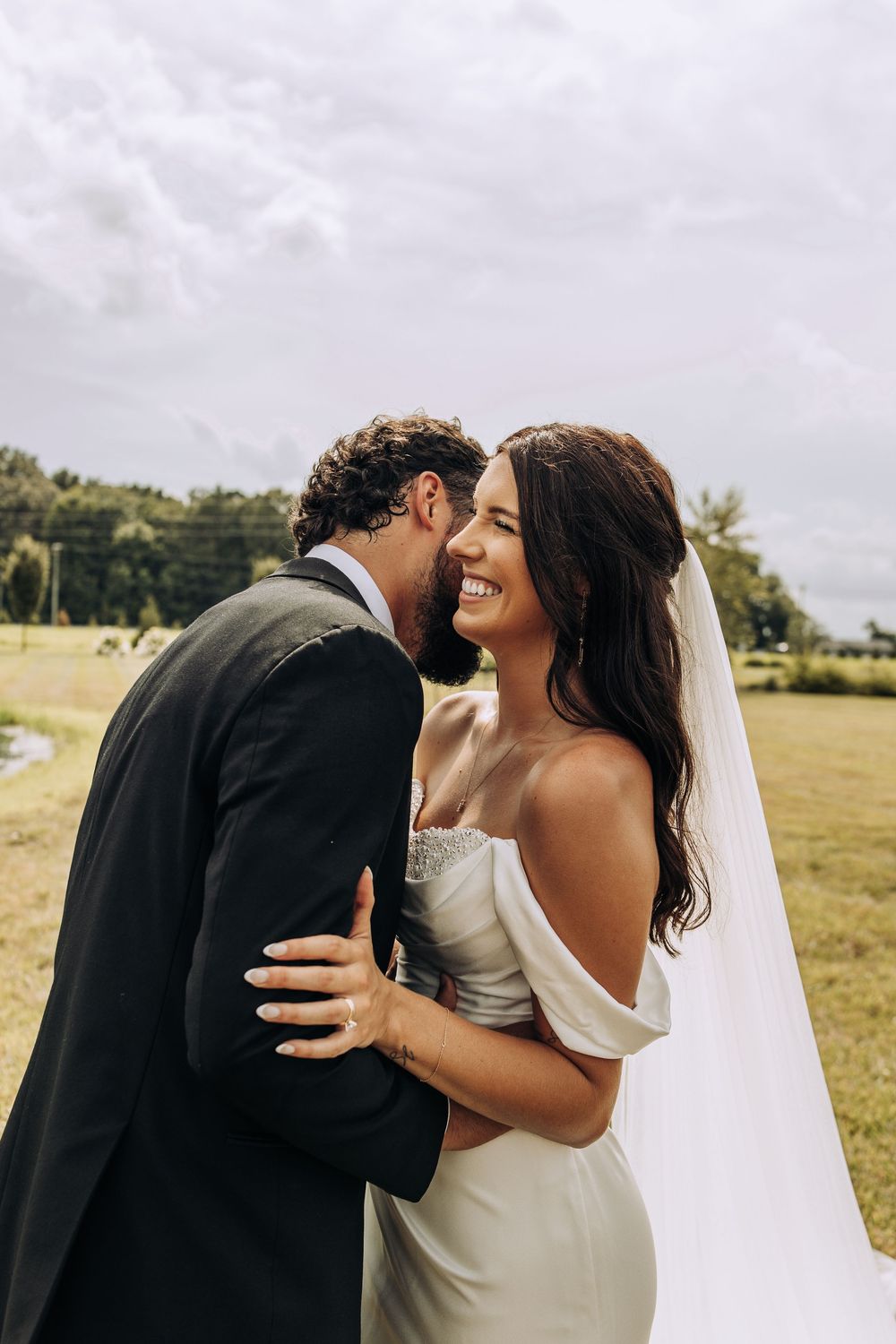 Smiling bride in off-shoulder gown with veil embraces groom in black suit outdoors.