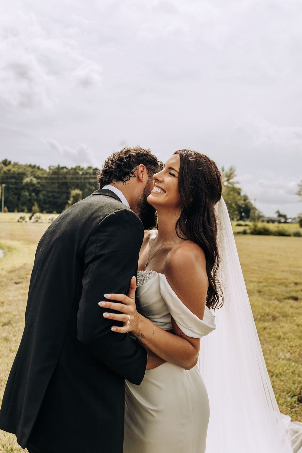 Radiant bride laughs joyfully as groom whispers in her ear during outdoor wedding portraits.