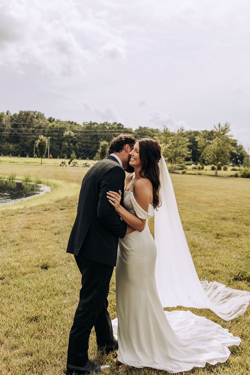 Bride in elegant white gown with cathedral veil shares intimate moment with groom in field.