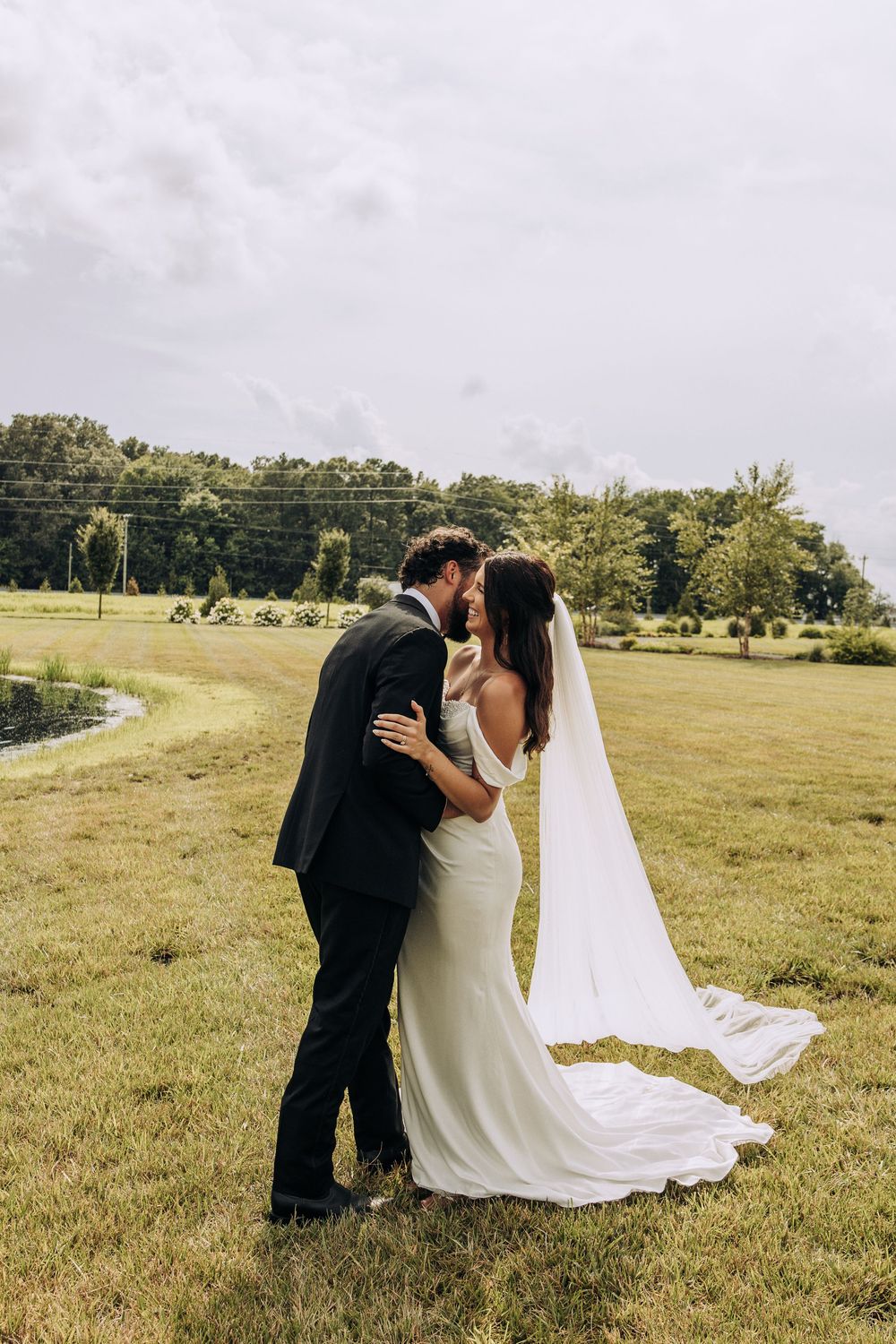 Wedding couple shares a kiss in green countryside setting with long bridal veil trailing.