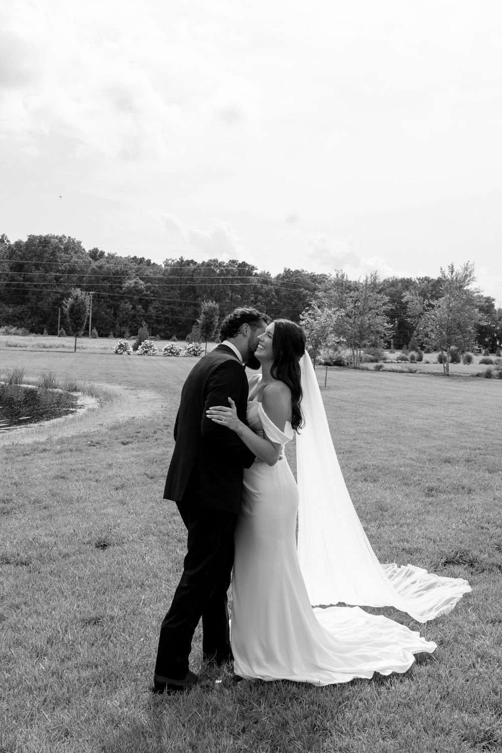Black and white romantic portrait of newlyweds kissing in open field with flowing bridal veil.