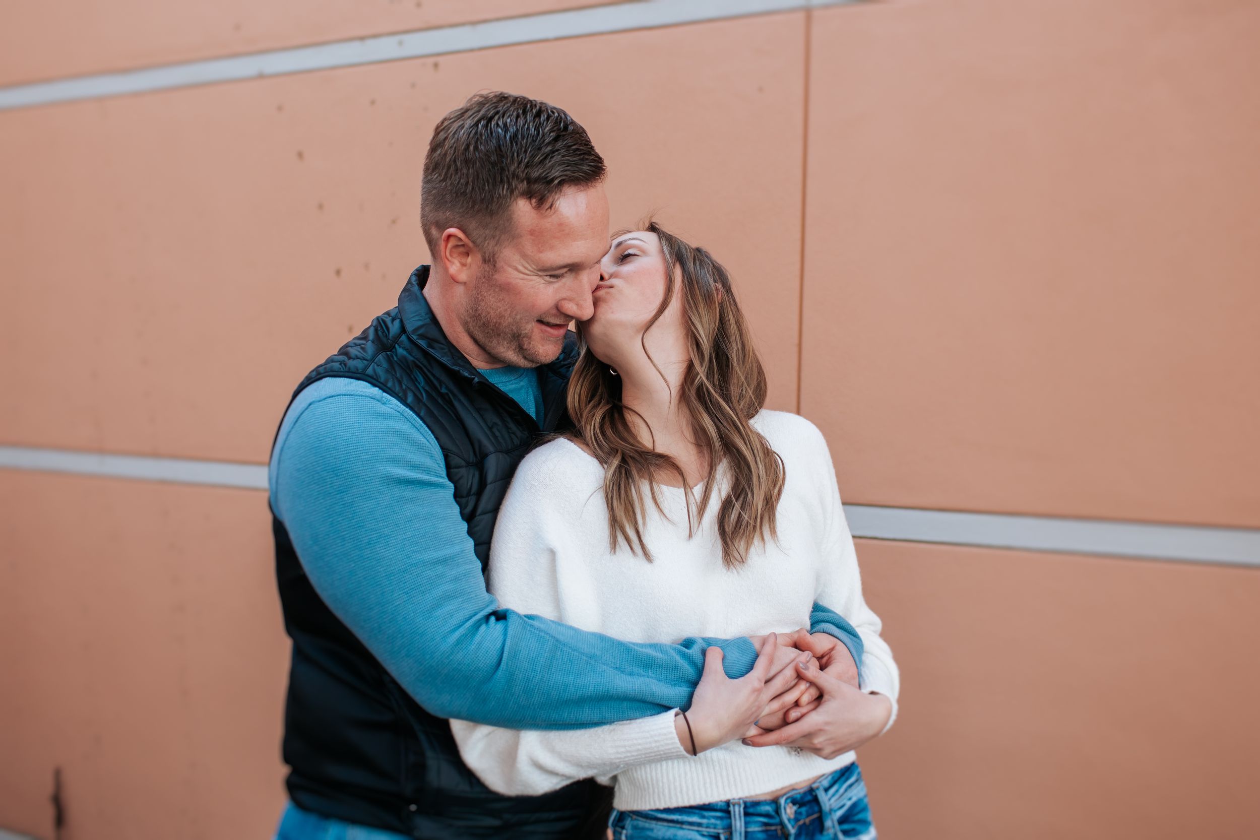 A couple in matching blue and white outfits embrace against a peach-colored wall in a romantic photoshoot.