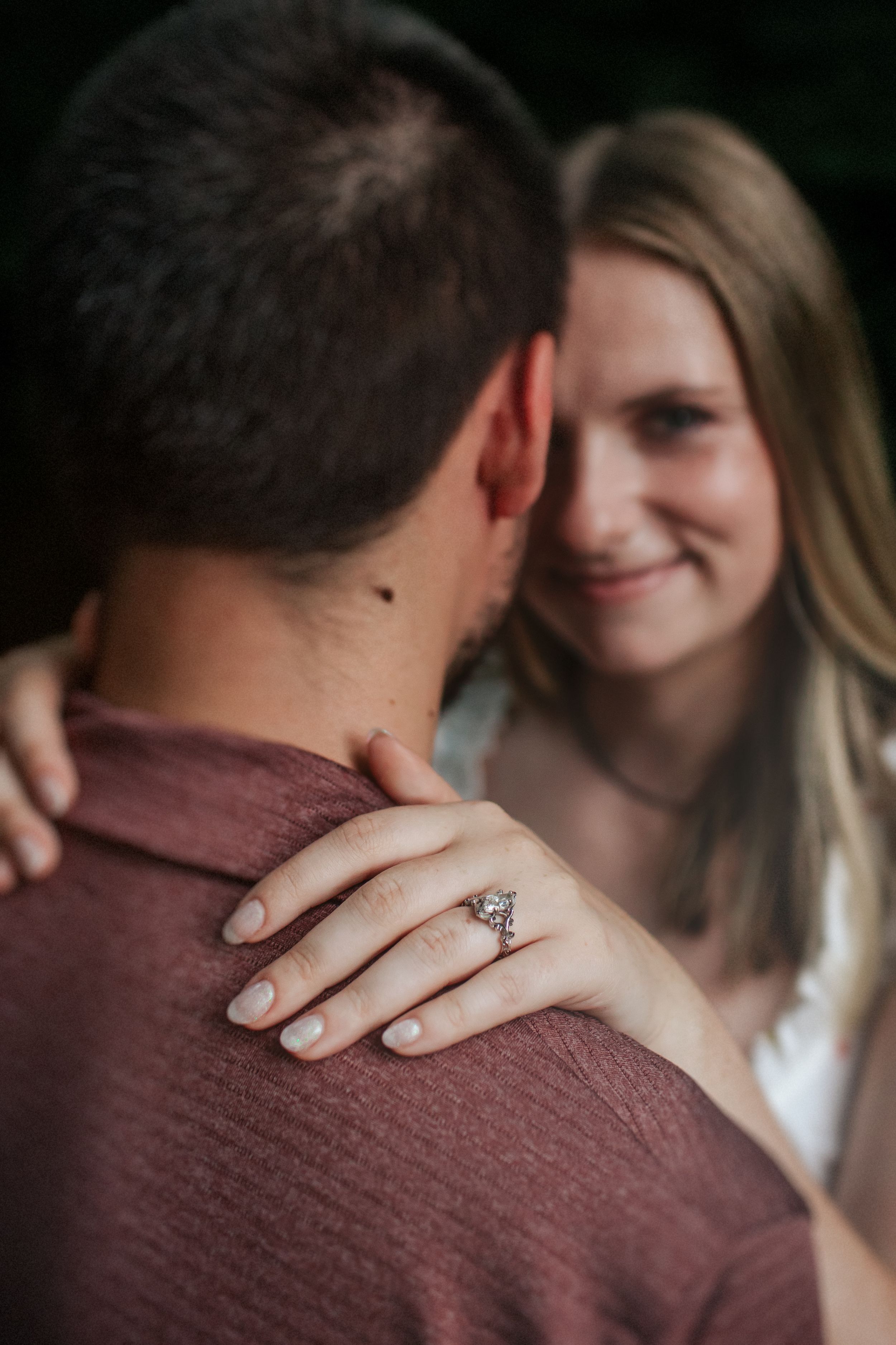 Close-up engagement ring shot with couple embracing in burgundy sweater showing romantic moment.