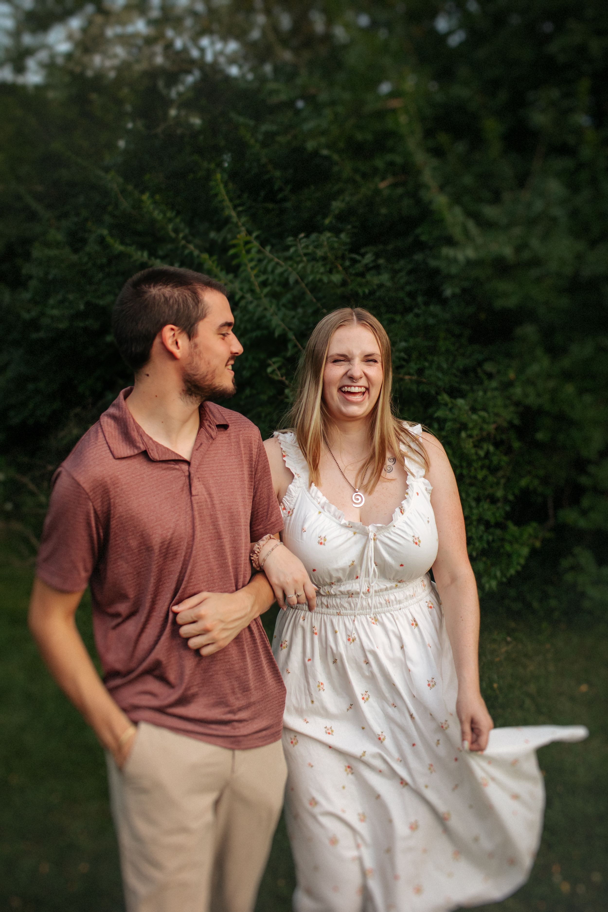 A couple shares a joyful moment outdoors, with her wearing a white dress and him in a burgundy polo and khakis.