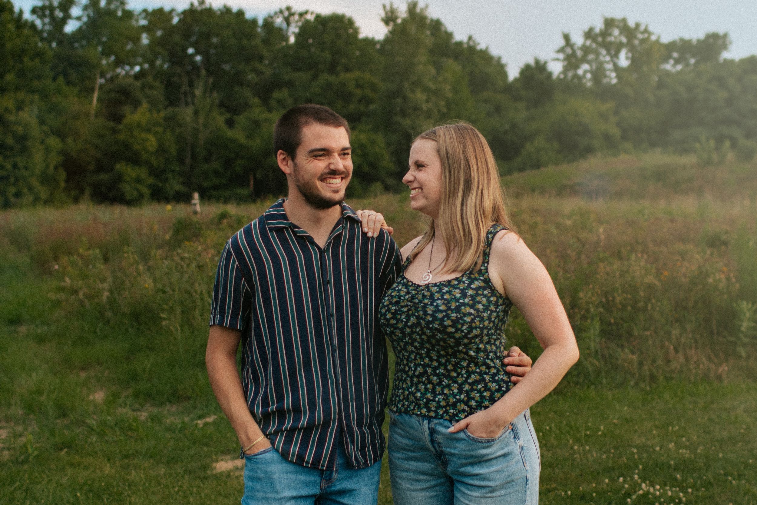 A couple sits together in a grassy meadow during a sunny outdoor engagement photo session.