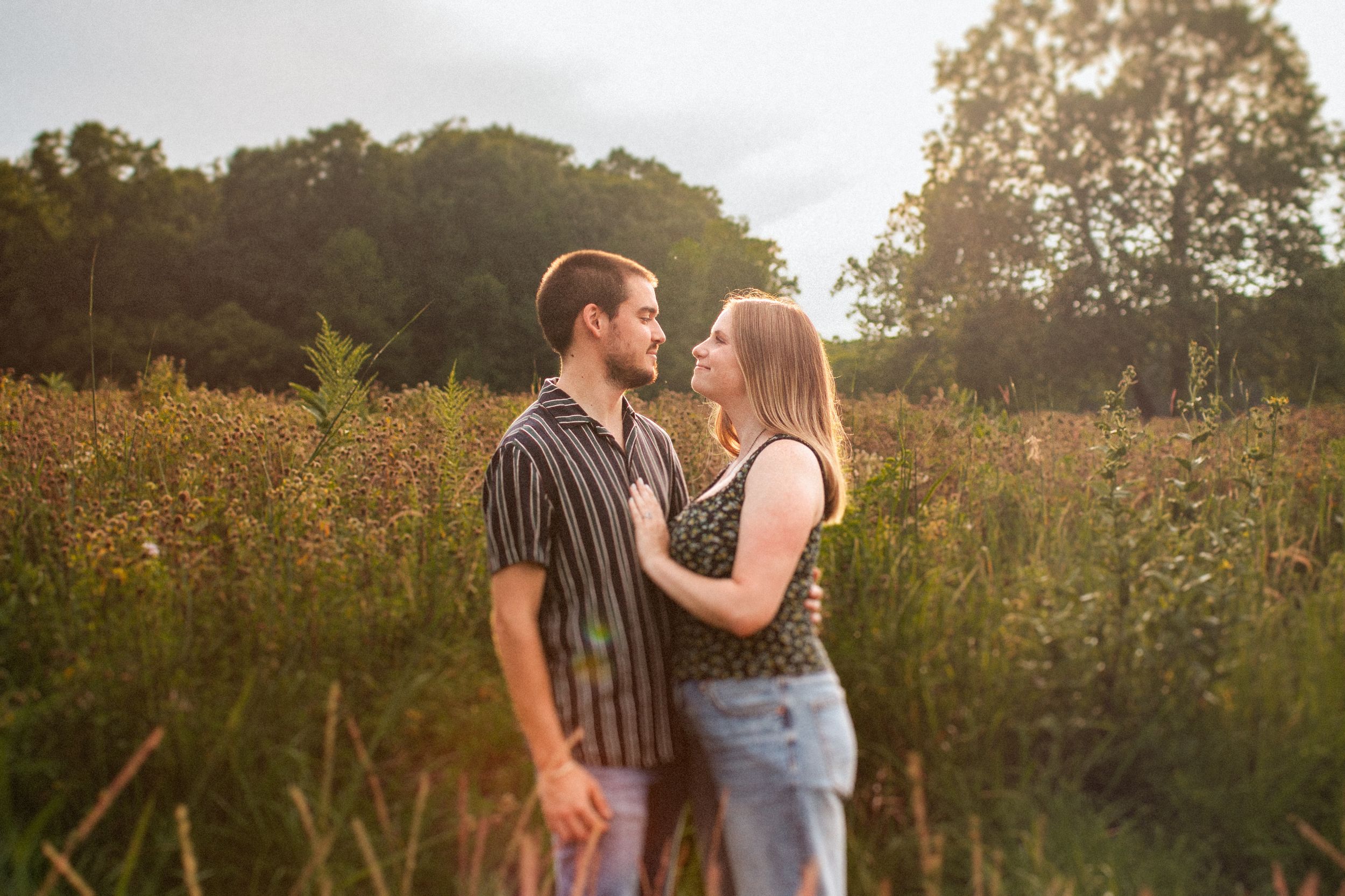 Couple embracing in meadow with warm sunlight filtering through trees during outdoor photo session.