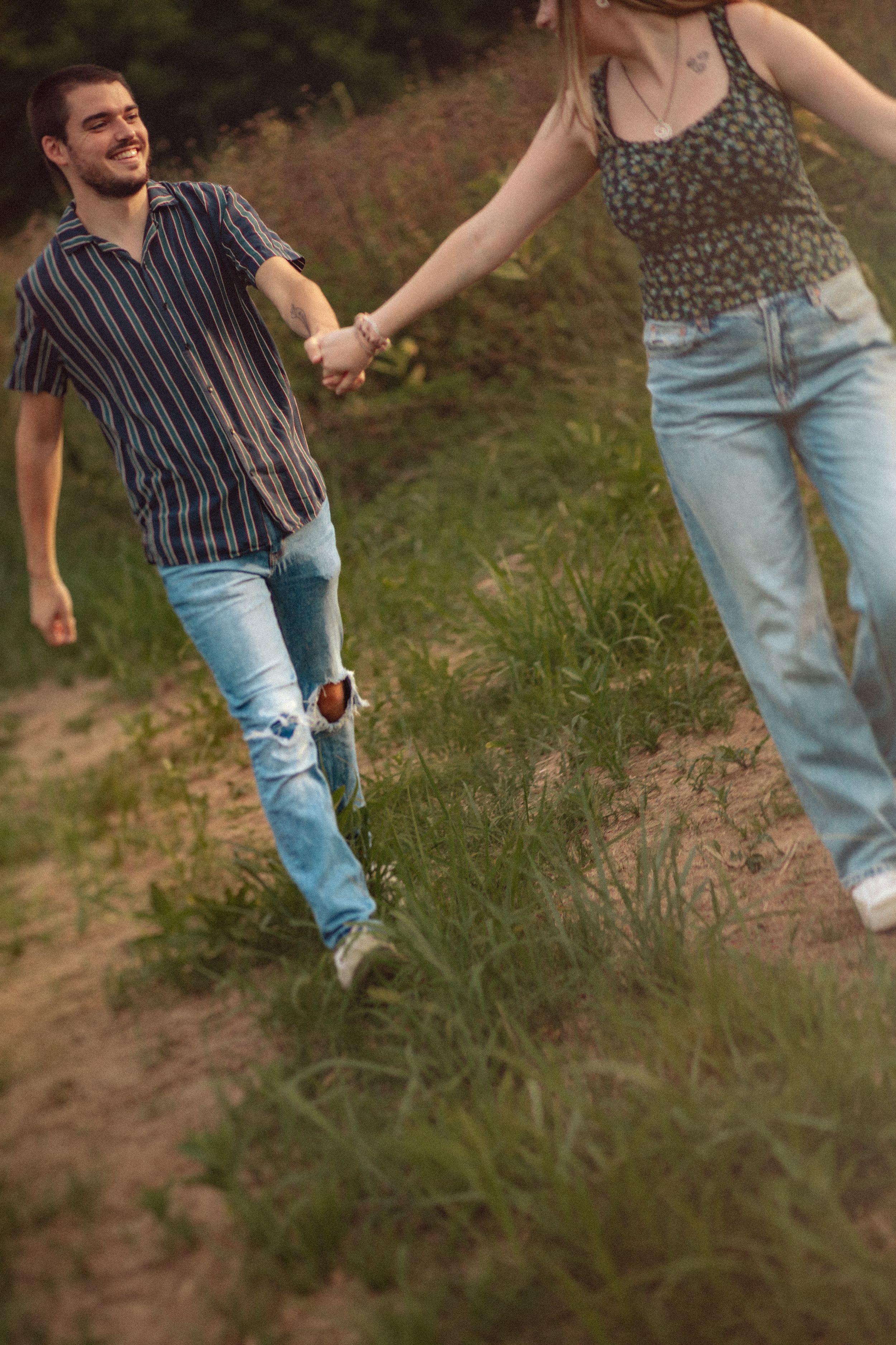 A couple walking together on a dirt path through grassy terrain while holding hands.