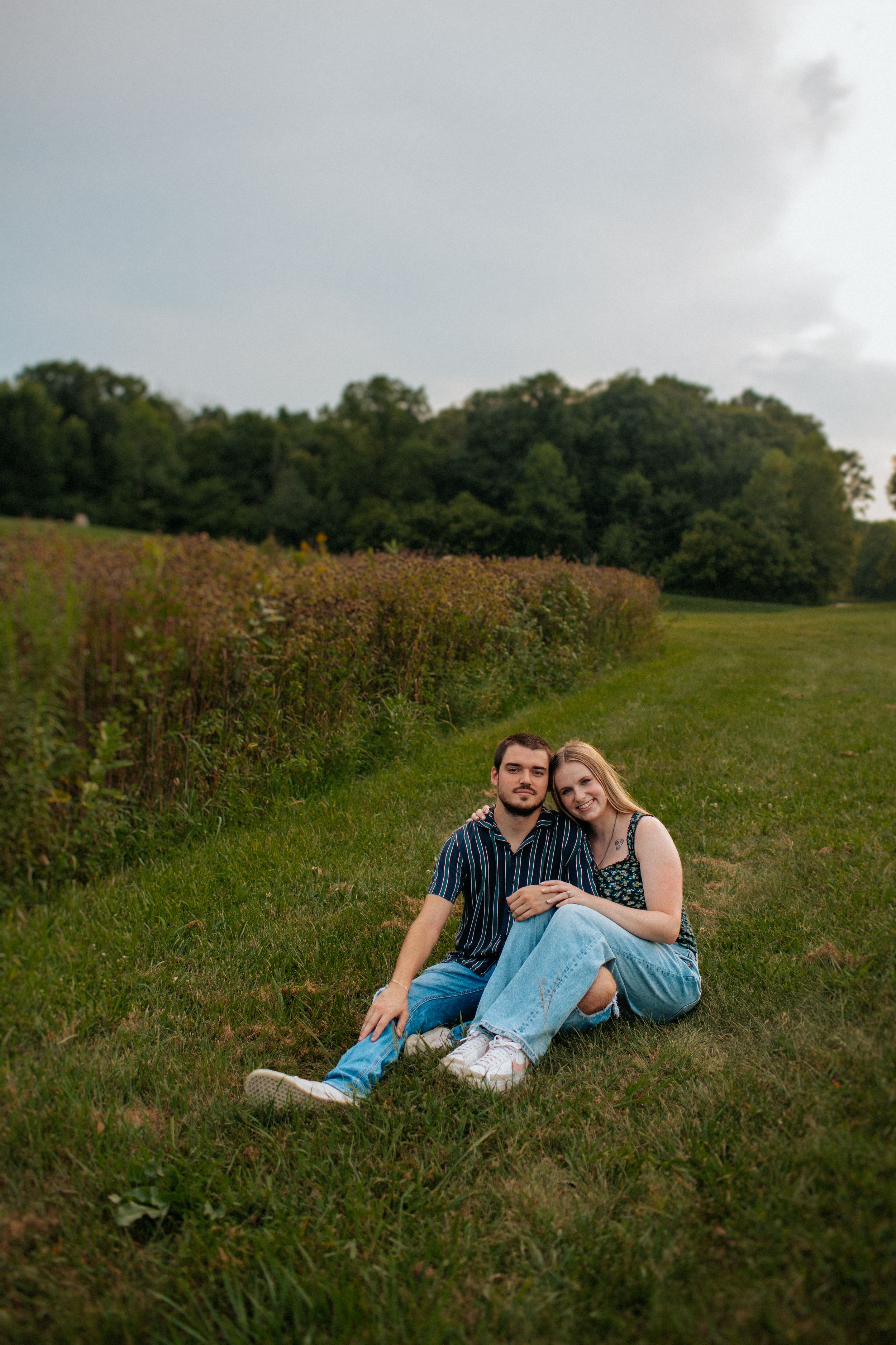 A couple relaxes together on grassy ground beside a wildflower field on a summer evening.